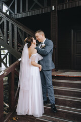 Fototapeta premium Stylish, young groom in a gray suit gently hugs a beautiful brunette bride in a white dress on the stairs, against the background of a wooden house. Wedding portrait of smiling newlyweds.
