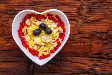 Puff salad with chicken breast and pineapple, garnished with pomegranate in a heart salad bowl on a wooden table.