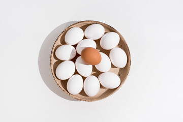 Un huevo de gallina marrón sobre una docena de huevos blancos en un plato rústico de color beige...