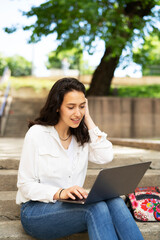 Young businesswoman sitting on steps outdoors and working on laptop. Beautiful girl learning in the park