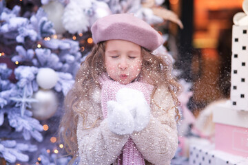 Portrait of little happy kid girl, beautiful positive cute blonde child is blowing snow from her hands, having fun at winter snowy day outdoors near Christmas tree in hat. Wintertime, happiness