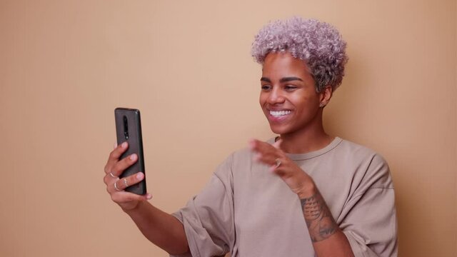 Young African American woman smiling looking at mobile phone and waving hand making online video call or recording appeal to subscribers standing on brown background. Blogging or podcasting concept