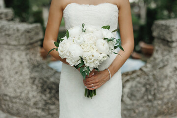Beautiful wedding bouquet of white peonies in hands of the bride Cropped photo Bride holding an elegant bouquet made of white flowers and greenery Stone wall background