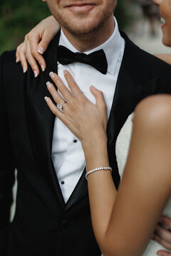 The gorgeous bride in white dress and stylish groom in a black suit with black bow tie gently hugging and smiling on background of green trees Sensual wedding couple embracing Newlyweds 