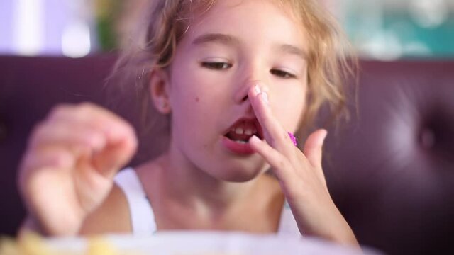 A 5-year-old girl is funny eating French fries in a cafe with her hands, chewing and making faces