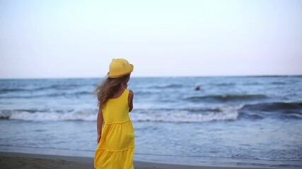 Girl in a yellow dress and hat with long hair fluttering from the breeze looks at the sea and waves, stands with her back to the frame. The end of the vacation, farewell to the sea, summer, nostalgia