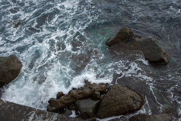 Sea stones top view. Aerial view of the grey seething sea surf. Big foaming waves break on the shore. Neutral natural background for the design. Drone view of the beach huge rocks. Tropical climate