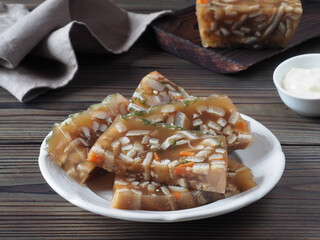 Pieces of meat jelly with pork skin, vegetables and herbs in a plate, horseradish sauce, napkin on a dark wooden table, flat layout, closeup. Traditional natural snack with jellied meat broth