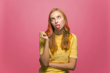 Funny young girl grimacing crossing eyes showing tongue, making foolish face expression, fooling on pink background