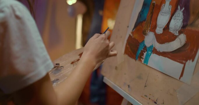 Close-up, the artist's hand picks up paint from a can and spreads it with a brush on a palette. Woman's hand paints a picture on an easel. Creative studio of the artist, dim lights in the studio. 4k