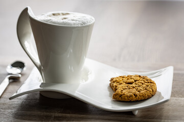 Modern cappuccino mug filled with aerated milk and a shot of espresso. Bitten oat meal cookie placed on the saucer.