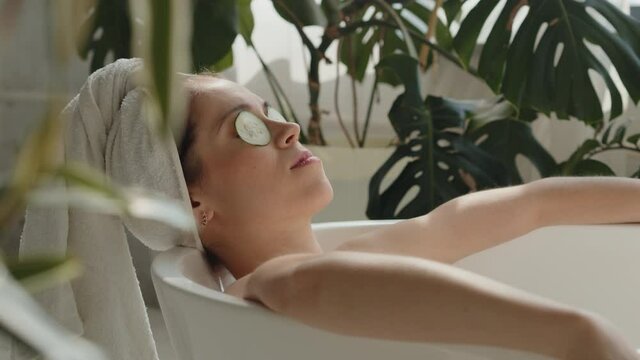 Pano shot of relaxed bautiful caucasian young woman lying in spa bath with cucumber on eyes and white towel on head, eyes closed. Relaxing spa wellness treatments in eco design bathroom