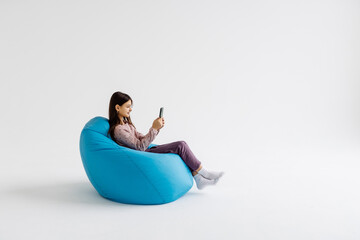 Little beautiful girl sitting on a beanbag holds mobile playing isolated on a white background.