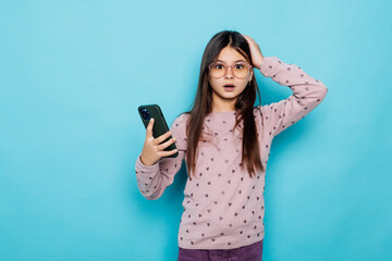 Naklejka premium Caucasian kid girl against blue wall hold hand modern technology use touch face palm astonished impressed scream wow omg