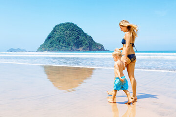 Happy family - mother, baby son have fun, walk by water pool along sea surf on sand beach. Red...