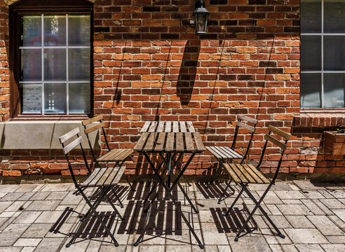 Weathered wood and iron bistro table and chairs sits on stone tiles in front of a rustic red brick wall with windows on a sunny day - Powered by Adobe