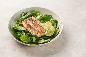 A plate with fresh green raw spinach and fried wild salmon, lime slices on grey concrete surface