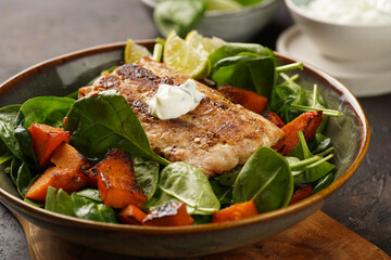 A plate with fresh green raw spinach and fried wild salmon, pumkin pieces, lime slices, cream cheese sauce, red chilli pepper on brown surface