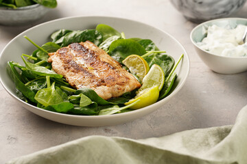 A plate with fresh green raw spinach and fried wild salmon, lime slices, cream cheese sauce on grey concrete surface