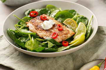 A plate with fresh green raw spinach and fried wild salmon, lime slices, cream cheese sauce, red chilli pepper on grey concrete surface