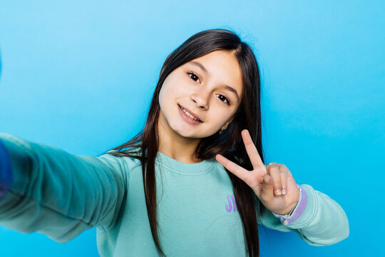 Portrait of happy friendly caucasian little girl over blue background taking selfie with peace gesture