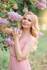 a girl with blonde hair in a pink dress stands next to a branch of lilac