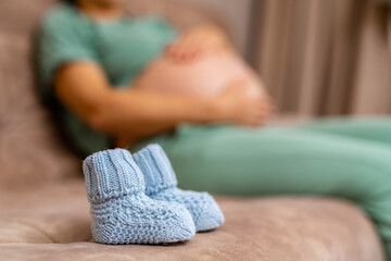 Little knitted socks for future child. Blue baby booties on sofa at the blur background of a pregnant woman resting on the sofa