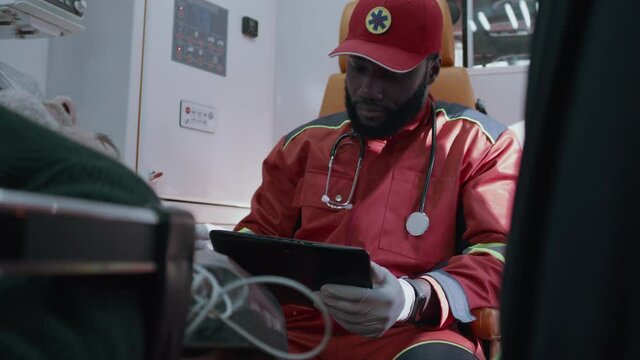 African American doctor in red uniform checking vital signs monitor of patient and entering medical data into tablet while riding emergency medicine vehicle to hospital