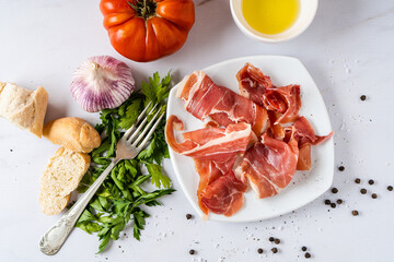 Raw or Spanish serrano ham on a white plate on a table with organic tomato and vegetables.