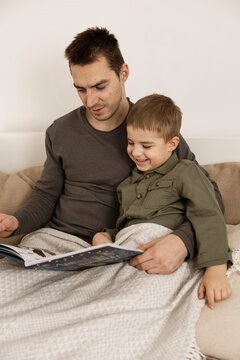 Father and son reading a book on the bed at home. Young attractive man and little boy resting in bedroom. Natural earth colors. Cozy environment. Father reads a fairy tale for his child.