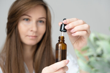 Young caucasian long haired brunette woman defocused doing body,skin care beauty procedures at home,holding glass bottle with serum,oil,bio natural cosmetics for hair or face.Selective focus