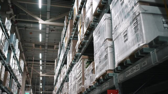 Rows of high metal storage racks with boxes of stuff items standing on shelves and wrapped in polyethylene in big warehouse of mall