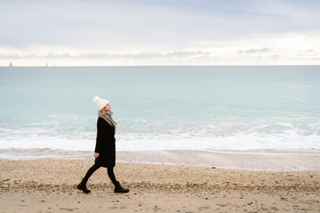 une femme blonde habillée avec un long manteau et un bonnet blanc , marche le long d'une plage