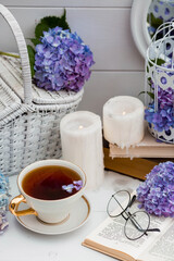 A visual for content. Still life in vintage style. A mug with a drink, an old book, a cage, candles and hydrangea flowers in the garden on a white wooden table. The concept of a tea ceremony.