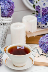 A visual for content. Still life in vintage style. A mug with a drink, an old book, a cage, candles and hydrangea flowers in the garden on a white wooden table. The concept of a tea ceremony.