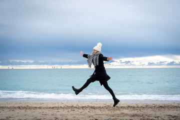 une jeune femme avec un bonnet , en hiver , saute de joie sur une plage