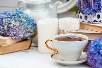 A visual for content. Still life in vintage style. A mug with a drink, an old book, a cage, candles and hydrangea flowers in the garden on a white wooden table. The concept of a tea ceremony.