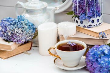 A visual for content. Still life in vintage style. A mug with a drink, an old book, a cage, candles and hydrangea flowers in the garden on a white wooden table. The concept of a tea ceremony.