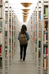 Figura feminina de costas andando por corredor de livros de uma biblioteca. 