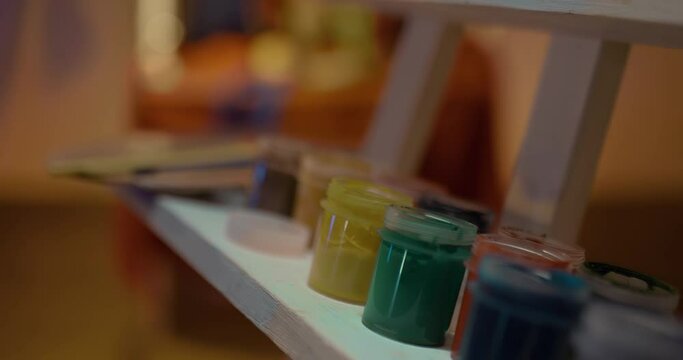 Close-up, cans of different colors are in a row. Brushes, an eraser, and a glass of water are on the easel. Artist workplace, light art class. 4k, ProRes