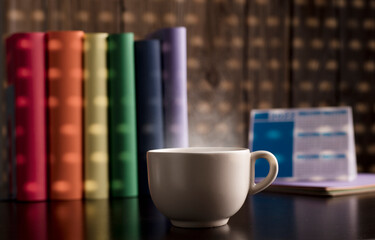 cup of coffee or tea with hot steam, books with lgtbi flag colors, on the desk, dawn light comes through the blind