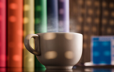 cup of coffee or tea with hot steam, books with lgtbi flag colors, on the desk, dawn light comes through the blind