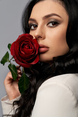 Charming caucasian woman holding red rose near plump lips and looking at camera over grey background. Young woman with trendy makeup and dark wavy hair posing in studio.