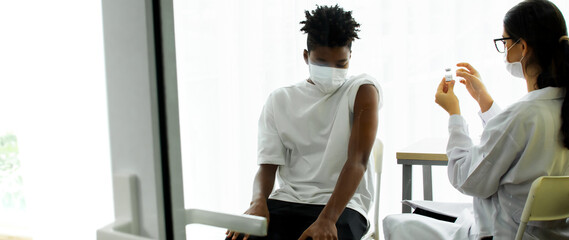Banner throgh window of African teen boy wearing face mask, getting vaccination to protect or prevent virus, getting vaccine injection while caucasian doctor preparing syringe at hospital.