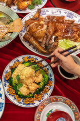 A hand holding chopsticks tries to pick up pieces of walnut shrimp on a table filled with different Chinese food dishes for Lunar New Year. 