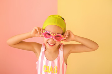 A child in a swimsuit and glasses and a swimming cap smiles sweetly looking at the camera. Girl swimmer Pink and yellow studio background.