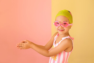 A child swimmer in swimming goggles stands with his hands folded in a pose for swimming. A girl in a beanie and striped swimsuit for swimming in the pool