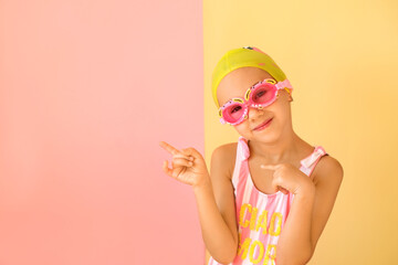 Girl swimmer in googles and swimming cap pointing with fingers aside at copy space on an pink studio background. Fashionable children's swimming goggles in the shape of a crab - Powered by Adobe