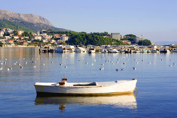 Obraz premium Small rustic boat and flock of seagulls in the sea. Podstrana, small touristic town near Split, Croatia, is seen in the background. 