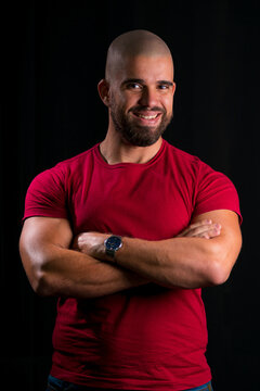 Retrato de homem careca jovem em est&uacute;dio com fundo preto. homem vestindo blusa vermelha com bra&ccedil;os cruzados, olhando para a camera e sorrindo.